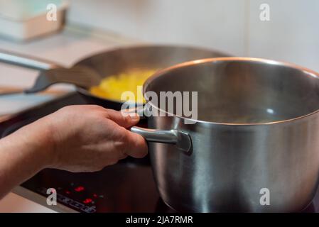 Main d'une femme tenant une casserole qui fait bouillir sur une table de cuisson en céramique. Banque D'Images