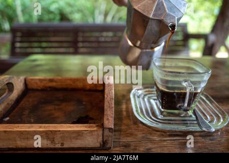 Versez à la main le café noir de la marmite moka dans une tasse de café sur une table en bois et un arrière-plan flou de la scène naturelle. Banque D'Images