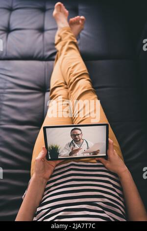 Femme couchée sur un canapé et parlant avec un médecin en ligne à l'aide d'une tablette numérique. Concept de télémédecine. Banque D'Images