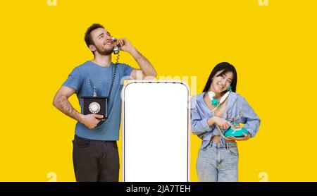 Fille asiatique et homme caucasien dans l'amour parlant sur les téléphones vintage adossé sur un énorme smartphone avec écran blanc, regardant l'un l'autre isolé sur fond jaune. Positionnement du produit. Banque D'Images