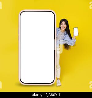 Adorable asiatique jeune fille excitée peeking dehors grand téléphone cellulaire vertical géant tenant un plus petit dans la main avec écran blanc vierge isolé sur fond jaune. Offre exceptionnelle. Positionnement du produit. Copier l'espace. Banque D'Images