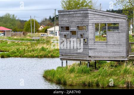 Un rideau de canard en bois surélevé à côté d'un étang dans un parc. Beaucoup de trous rectangulaires à regarder à travers. Quartier résidentiel derrière, ciel couvert. Banque D'Images