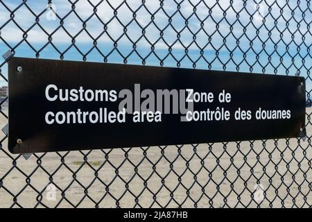 Une affiche bilingue de zone contrôlée par les douanes en anglais et en français sur une clôture à chaînette. Vue en angle avec ciel bleu au-dessus, parking en dessous. Banque D'Images