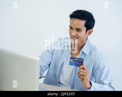 Joyeux homme asiatique souriant dans le magasin de chemise en denim en ligne, payant avec une carte de crédit dans sa main avec un ordinateur portable pour sa propre petite entreprise sur un W. Banque D'Images