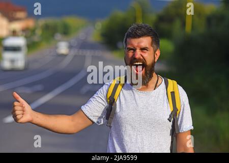 Arrêt automatique du déplacement. Taille basse essayez d'arrêter la voiture et le pouce vers le haut geste. Homme au visage joyeux et à la barbe voyageant en randonnée avec la route en arrière-plan Banque D'Images