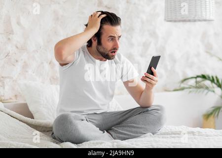 Jeune homme choqué assis sur le lit avec un smartphone, en touchant la tête Banque D'Images