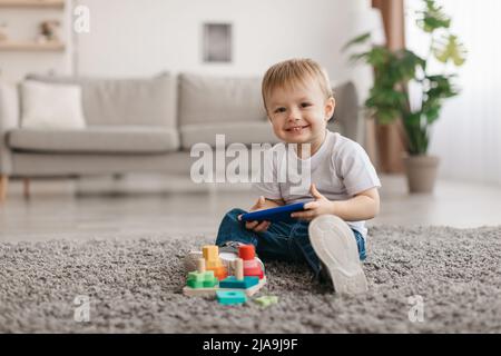 Portrait d'un adorable petit garçon tenant un smartphone, assis sur la moquette, regardant et souriant l'appareil photo, espace de copie Banque D'Images
