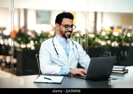 Gai jeune médecin islamique en lunettes, manteau blanc travaille à l'ordinateur dans l'intérieur moderne de bureau de clinique Banque D'Images
