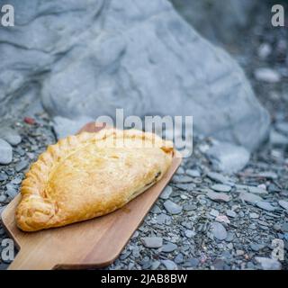 Pasty cornouaillais sur une assiette en bois sur une plage rocheuse Banque D'Images