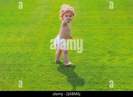 Bébé marchant pieds nus sur de l'herbe verte dans une soirée ensoleillée d'été. Petit garçon jouant dans le parc d'été dans un pantalon de couches. Banque D'Images