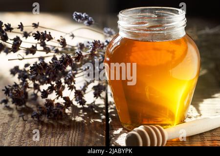 Miel dans un pot en verre, une cuillère pour le miel, fleurs de lavande sur une vieille table en bois. Arrière-plan sombre. Lumière naturelle de la fenêtre. Banque D'Images