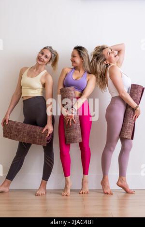 trois jeunes femmes souriantes parlant tenant le tapis de yoga dans le sport vêtements en studio blanc, professeur de yoga, entraînement Banque D'Images