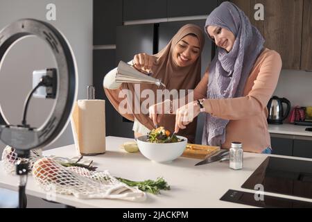 Deux bûcherons musulmans souriant tout en versant de l'huile d'olive dans une salade dans la cuisine. Banque D'Images