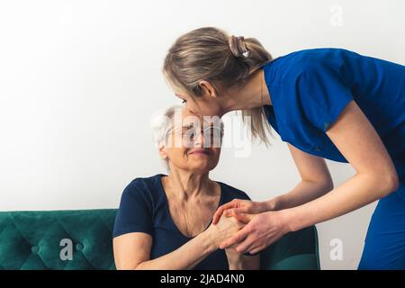 Une femme MD dans son 20s portant des gommages bleus se penchant vers une femme âgée assise sur un canapé, l'embrassant dans son front. Prise de vue en intérieur. Photo de haute qualité Banque D'Images