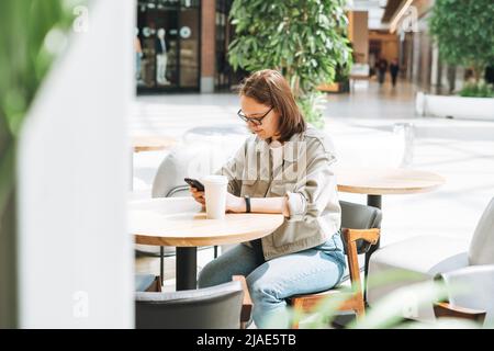 Jeune brune adolescente jeune fille étudiante en lunettes utilisant le téléphone mobile au café vert moderne, espace ouvert public Banque D'Images