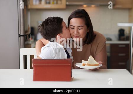 Adorable petit garçon embrassant la mère sur les joues après qu'elle lui a apporté des sandwiches pour le petit déjeuner Banque D'Images