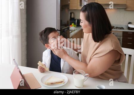 La mère qui coud les joues de son fils lorsqu'ils prennent le petit-déjeuner en cuisine Banque D'Images