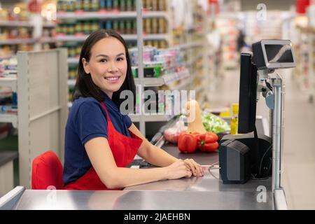 Femme gaie travaillant dans un supermarché assis à la caisse qui regarde à l'appareil photo souriant Banque D'Images