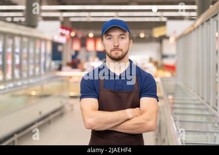 Portrait horizontal moyen d'un jeune homme moderne portant un uniforme de travail dans le magasin debout, bras croisés en regardant la caméra Banque D'Images