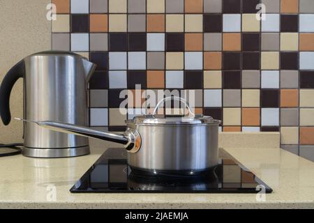 Une casserole en acier sur une cuisinière à induction, une bouilloire électrique en arrière-plan. Photo d'une cuisine avec carreaux de couleur sur le mur et comptoir en pierre Banque D'Images