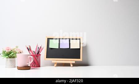 Tableau noir avec notes adhésives colorées, porte-crayon, tasse à café et pot à fleurs sur table blanche Banque D'Images