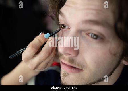 Portrait d'un jeune homme avec une barbe posant pour le maquillage, main femelle appliquant un eyeliner sur ses yeux Banque D'Images