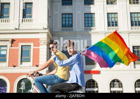 Jeunes hommes gay couple à vélo tenant gay fierté drapeau dans la ville regardant la caméra. concept lgbt Banque D'Images