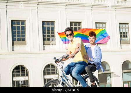 Jeunes hommes gay couple à vélo avec drapeau de fierté gay regardant la caméra à l'extérieur. concept lgbt Banque D'Images