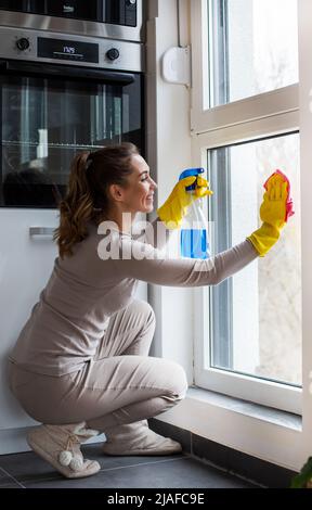 Une jeune femme en gants de protection en caoutchouc nettoie la fenêtre avec un chiffon en microfibre et tient le vaporisateur dans son autre main Banque D'Images