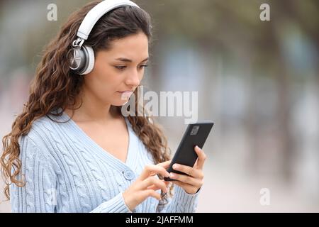 Femme sérieuse avec un casque sans fil écoutant de la musique vérifiant smartphone dans la rue Banque D'Images