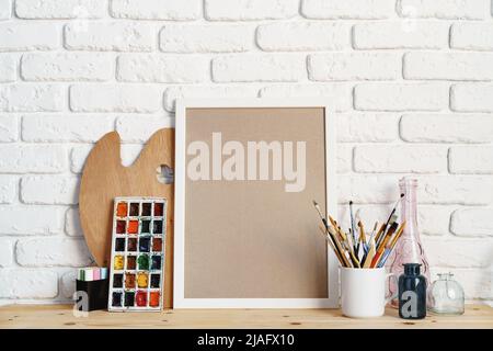 Cadre photo comme chevalet avec les outils de l'artiste sur une table en bois contre un mur en brique blanche Banque D'Images