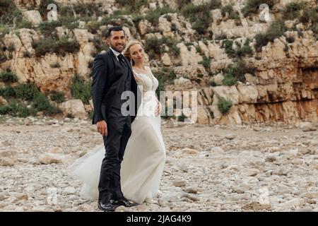 Portrait de jeune mariée blonde en robe blanche et marié brunet en costume souriant et marchant sur la côte rocheuse de la mer en Italie, paysage marin. Magnifique Banque D'Images