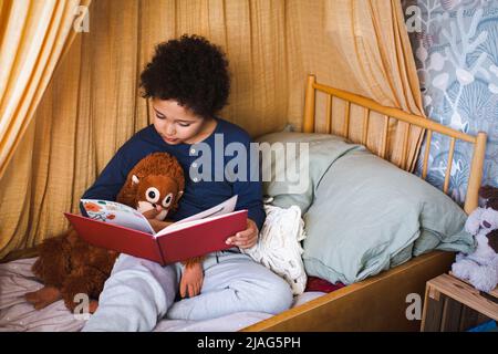 Garçon avec des jouets de trucs lisant le livre d'histoire tout en étant assis sur le lit à la maison Banque D'Images