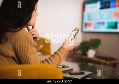 Vue latérale d'une jeune femme contrôlant la télévision via une application mobile à la maison Banque D'Images