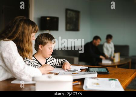 Garçon avec le syndrome de Down lisant le livre par la mère tout en faisant des devoirs à la table Banque D'Images