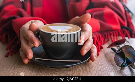 Une femme enveloppée dans une couverture à carreaux rouges tient une tasse chaude de café dans ses mains. Ambiance chaleureuse. Gros plan des mains d'âge moyen des femmes Banque D'Images