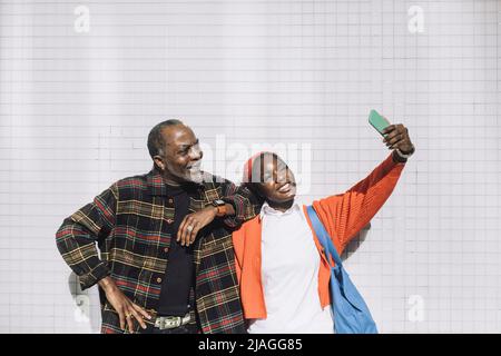 Une jeune femme heureuse prend le selfie avec son père à travers son smartphone tout en se tenant contre le mur Banque D'Images