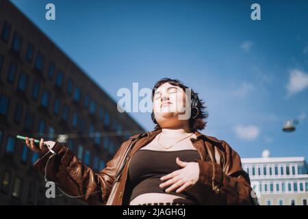 Jeune femme avec les yeux fermés écoutant de la musique par des écouteurs sans fil pendant la journée ensoleillée Banque D'Images