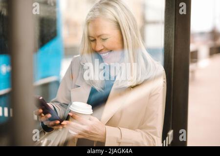 Bonne femme d'affaires utilisant un smartphone tenant une tasse jetable tout en s'appuyant sur le verre à l'arrêt de bus Banque D'Images