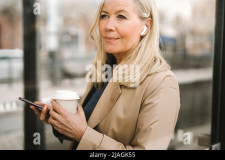 Femme d'affaires contemplant tout en tenant un smartphone et une tasse jetable à l'arrêt de bus Banque D'Images