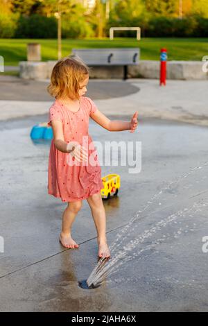 Petite fille jouant dans le parc avec des fontaines en été. Un petit enfant s'amuse à l'aire de jeux de l'aire de jeux en été Banque D'Images