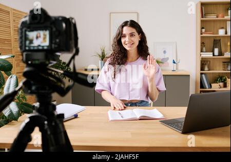 Jeune femme attrayante assise au bureau devant l'appareil photo sur trépied démarrage vidéo pour le blog avec salutation de ses disciples Banque D'Images