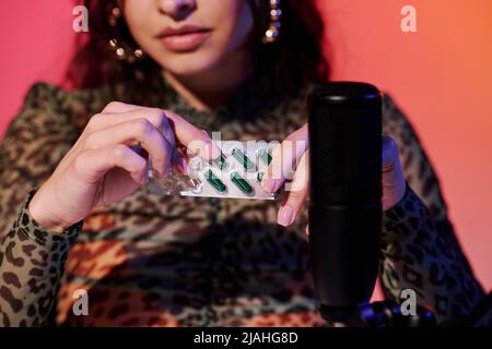 Jeune femme moderne assise en studio dans des sons d'enregistrement de lumière néon rose et jaune pour ASMR vlog en utilisant un blister pharmaceutique Banque D'Images