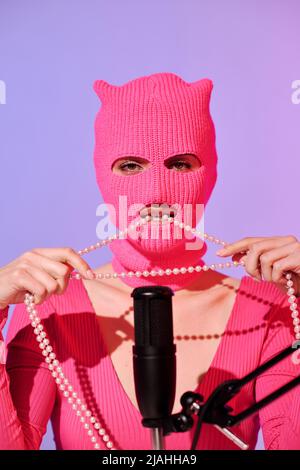 Jeune influenceur méconnaissable portant un élégant chandail rose et une balaclava assis devant le micro posé sur un collier de perles de mirage Banque D'Images