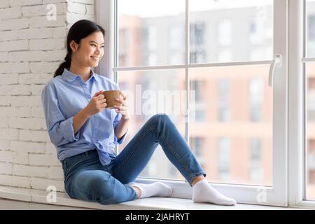 Positive jolie femme asiatique assise sur la fenêtre, buvant du café Banque D'Images