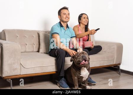 jeune couple regardant la télévision avec télécommande en main, pop-corn et leur chien sur un canapé Banque D'Images