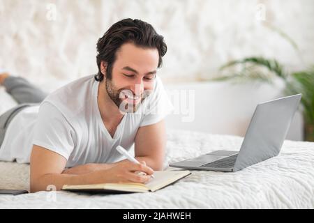 Jeune homme souriant, allongé sur le lit, utilisant un ordinateur portable, prenant des notes Banque D'Images