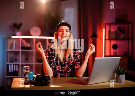 Femme agréable méditant avec les yeux fermés tout en s'asseyant au bureau avec un ordinateur portable moderne. Femme indépendante pratiquant le yoga pour soulager le stress au travail. Concept de personnes et de pensée positive. Banque D'Images