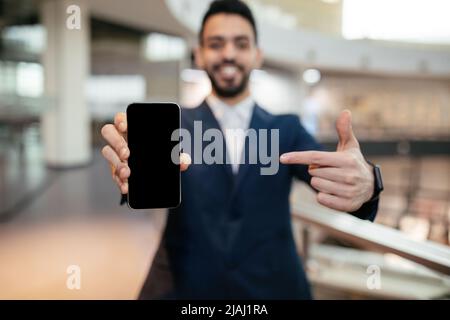 Un jeune directeur général musulman heureux avec une barbe en costume pointe le doigt sur le téléphone avec un écran vide au bureau Banque D'Images