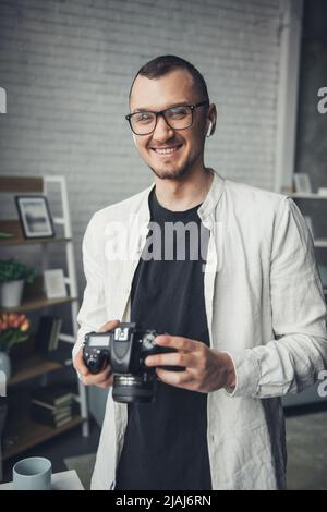 Portrait d'un beau photographe souriant tenant l'appareil photo, debout dans son studio. Le photographe professionnel règle l'appareil photo avant de prendre des photos. Banque D'Images
